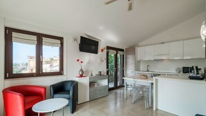 A modern living room with a red and black sofa, a small white table, and a kitchen area with white cabinets and a TV mounted on the wall.