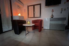 Ein kleiner, gut beleuchteter Raum mit einem Bett, einem Nachttisch, einem Sessel und einem an der Wand montierten Fernseher.