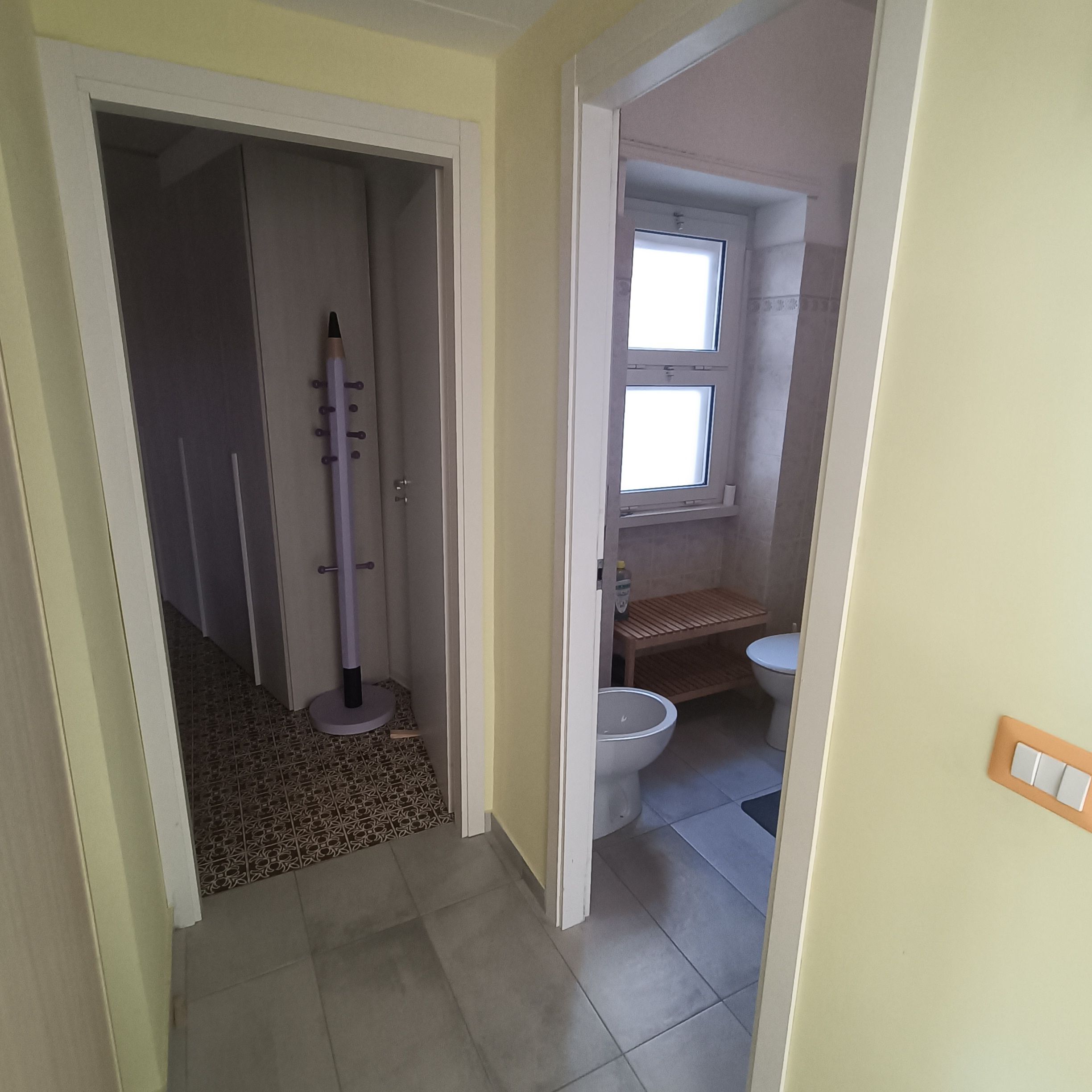 Un couloir étroit mène à une salle de bain équipée de toilettes, d’une petite fenêtre et d’une étagère en bois.