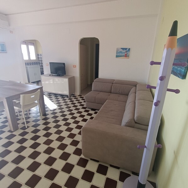 A living area with a checkered floor, a sofa, a TV on a cabinet, and a wall-mounted rack. - The beach is 20 meters away (Tarquinia Lido)