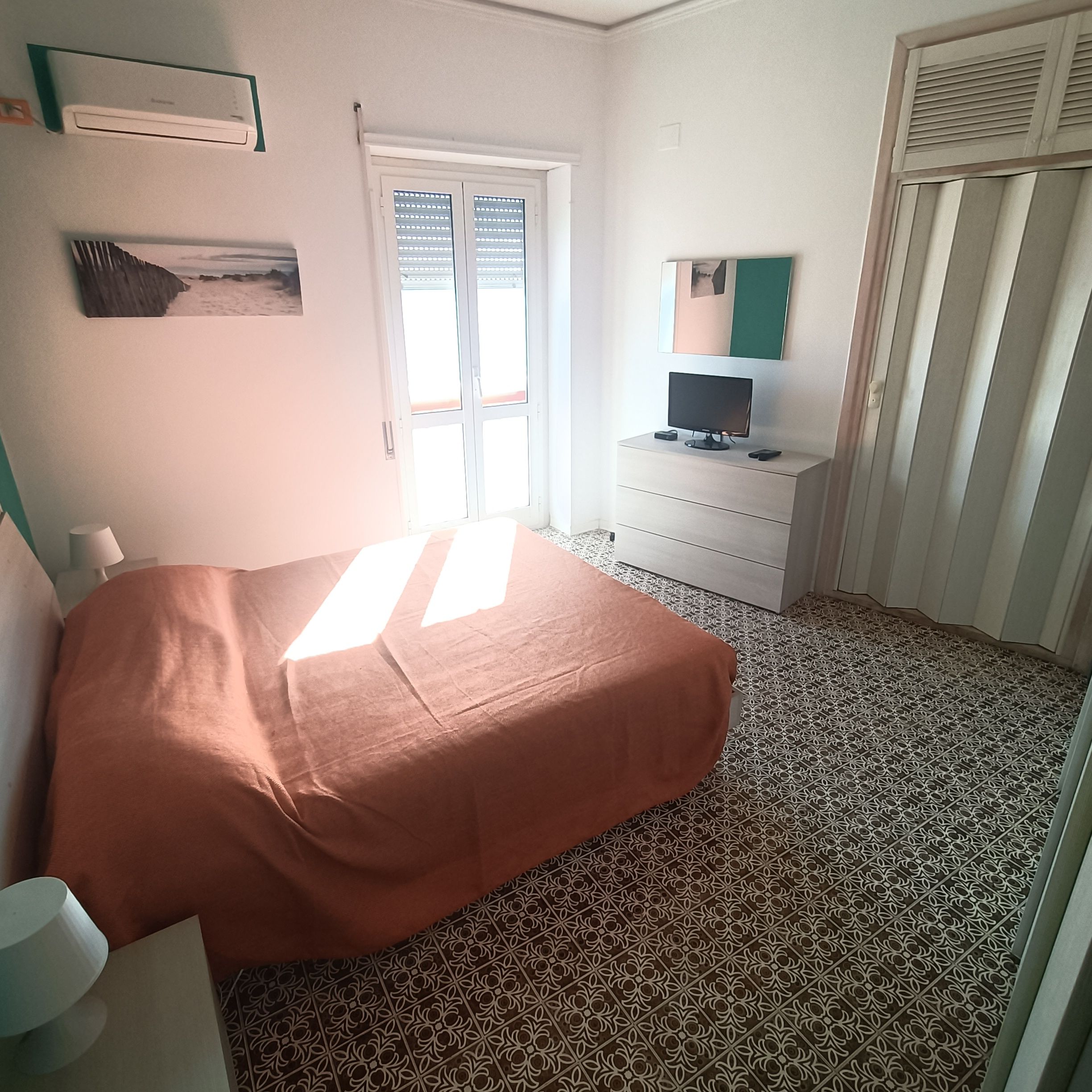 Une chambre à coucher comprenant un lit, une commode, un téléviseur et une fenêtre avec des stores.