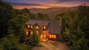 A two-story house with a stone chimney, surrounded by dense forest, and a gravel driveway.