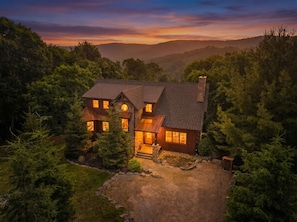 A two-story house with a stone chimney, surrounded by dense forest, and a gravel driveway.