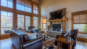 A spacious living room with large windows, a stone fireplace, leather furniture, and a mounted television.