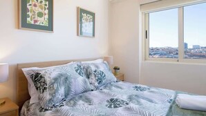 Una habitación de hotel con una cama, dos cuadros enmarcados en la pared, una mesita de noche con una lámpara y una ventana con vistas a la ciudad.