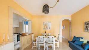 A dining area with a white table and chairs, a kitchen in the background, and a blue sofa with cushions.