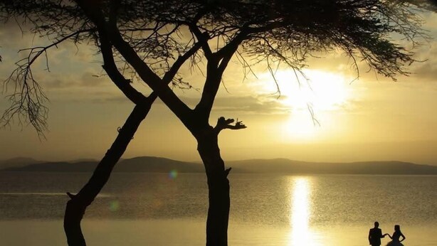 Silueta de una pareja tomándose de las manos contra un atardecer, con un lago tranquilo y colinas al fondo.