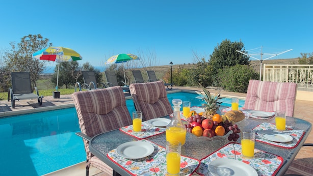 A poolside dining area with chairs, a table set for two with plates, glasses, and a fruit basket. - Luxury new villa on mountainside with private swimming pool & garden (Kathikas)