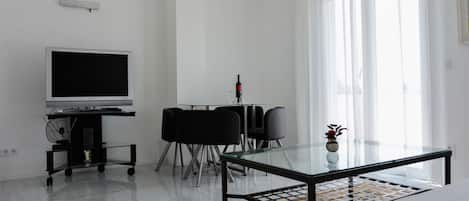 Una sala de estar moderna con un televisor de pantalla plana, una mesa de centro de vidrio, una zona de comedor con mesa y sillas, y ventanas grandes con cortinas translúcidas.