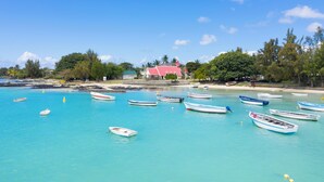 A coastal scene with several small boats anchored in clear turquoise waters, a sandy beach, and a prominent red-roofed building in the background.