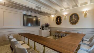 Un salón de conferencias con una larga mesa de madera, sillas blancas tapizadas, un televisor de pantalla plana y dos cuadros colgados en la pared.