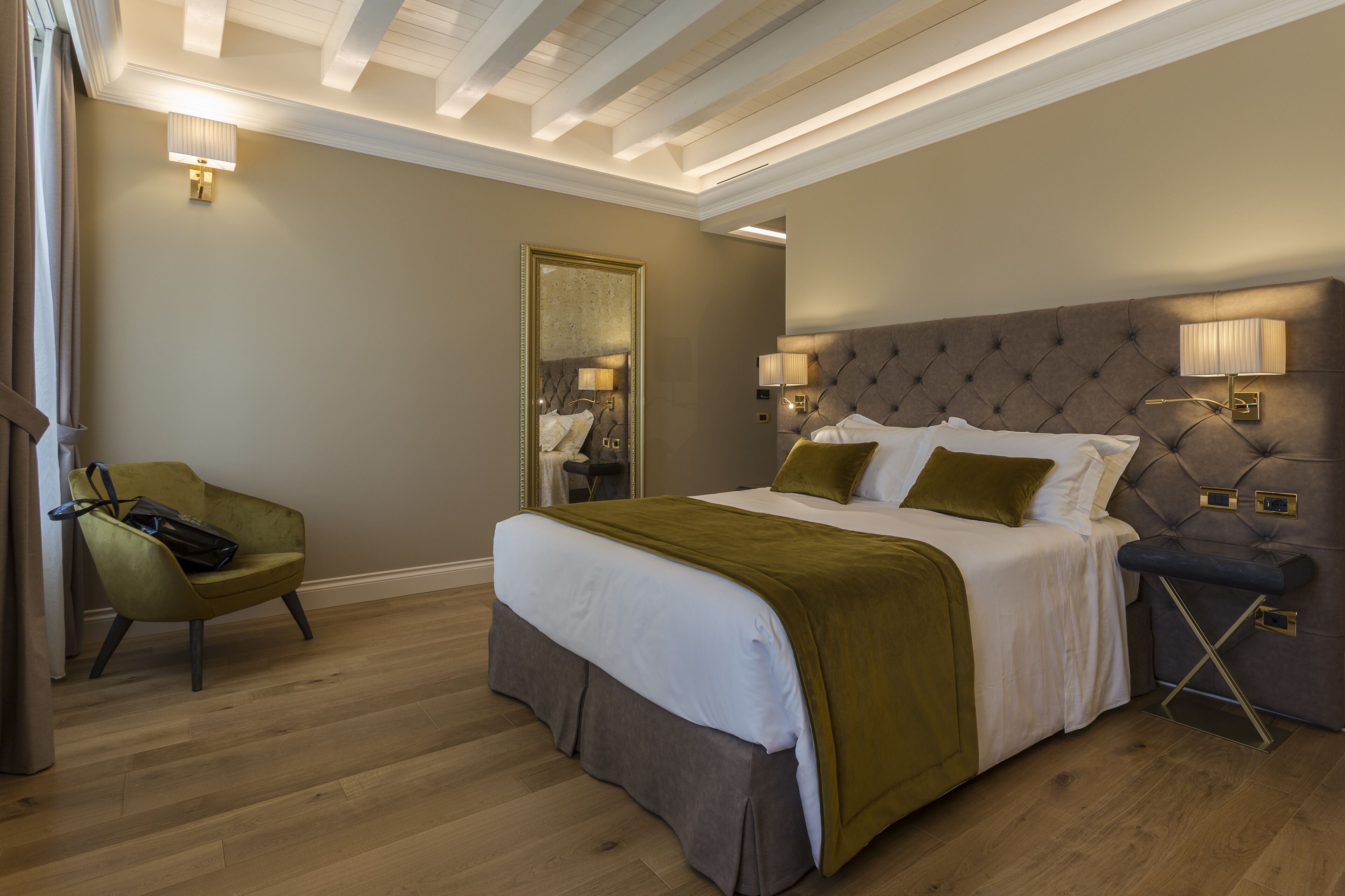 Een hotelkamer met een groot bed, een gestoffeerd hoofdeinde, bedlampen, een spiegel, een groene fauteuil en houten vloeren.
