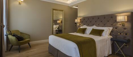 Habitación de hotel con una cama grande, cabecera capitonada, lámparas de noche, espejo, un sillón verde y suelo de madera.