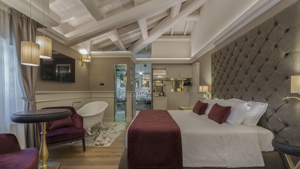 A modern hotel room with a large bed, a sitting area with a chair and table, a bathtub, and a well-lit ceiling with exposed beams.