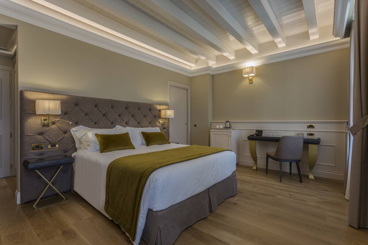 A bedroom with a tufted headboard, a bed with white linens and green accents, a desk with a chair, and a small kitchenette.