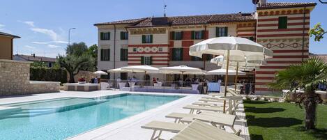 A swimming pool with lounge chairs and umbrellas, a stone wall, and a building with striped architecture in the background.