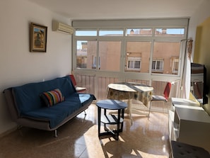 A room with a blue sofa, a small round table, and a chair. There is a window with a view of buildings outside.