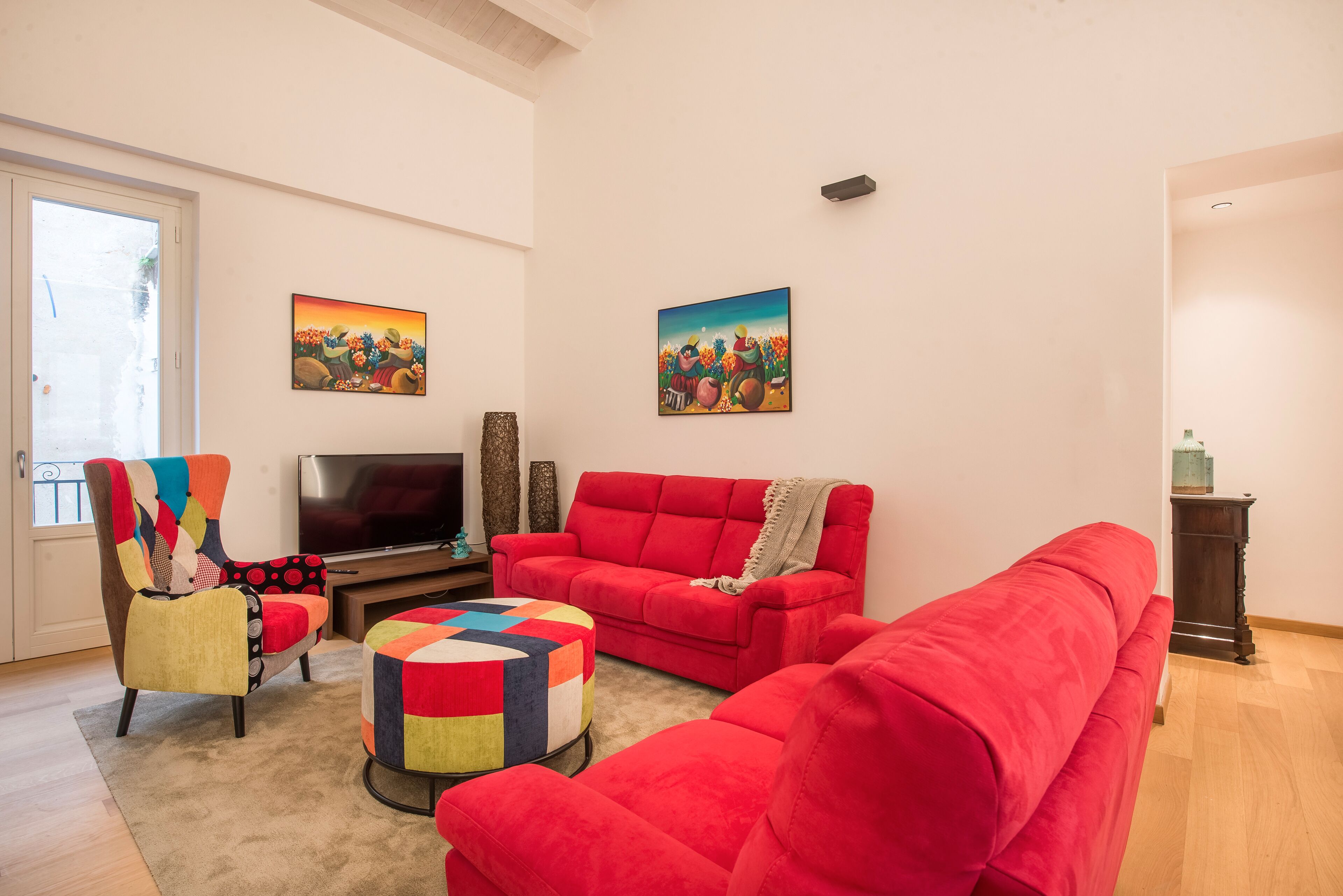A living room with a red sofa, a colorful armchair, and a TV. There are two paintings on the wall and a wooden floor.