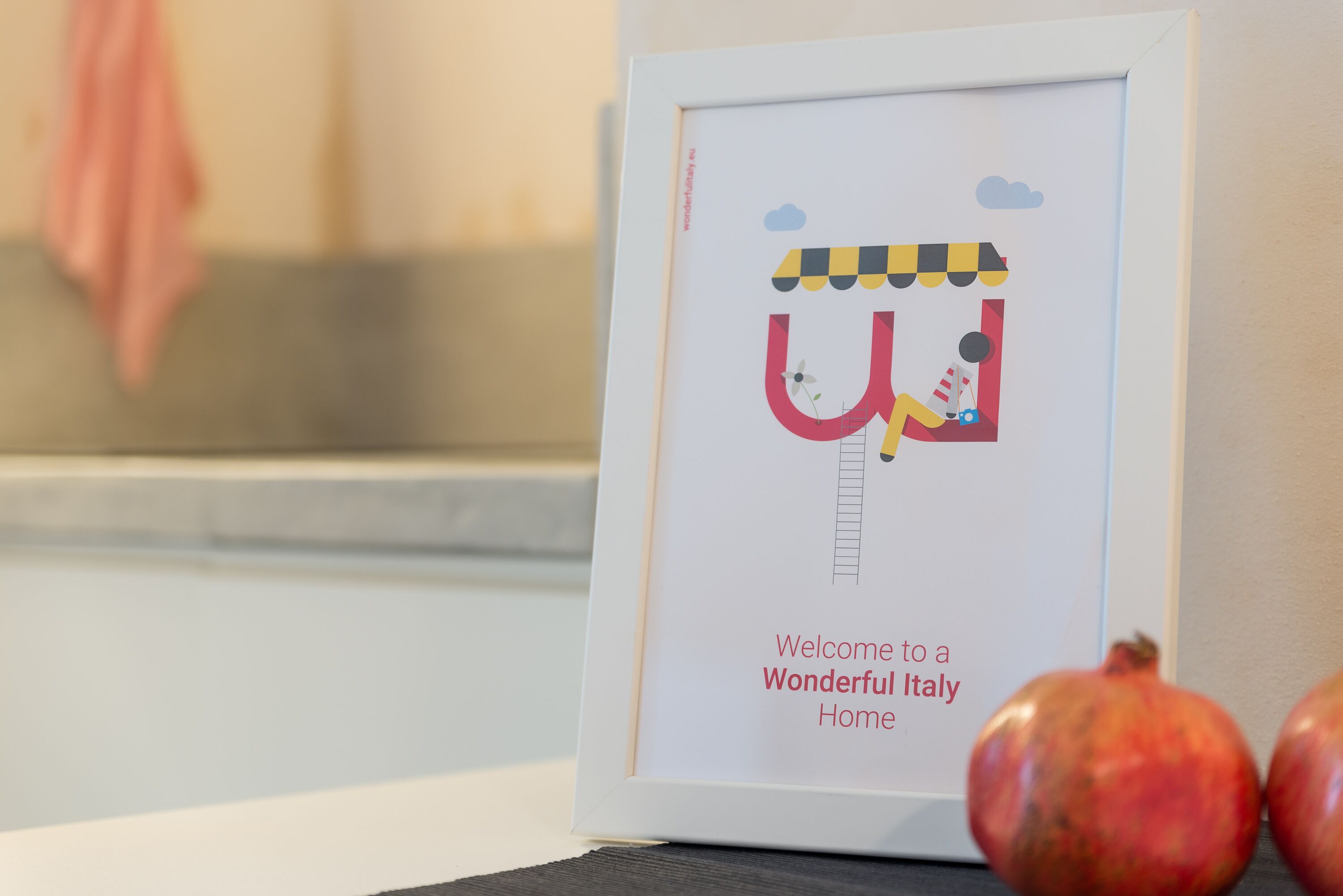 A framed welcome sign with a ladder and a character, placed on a table with pomegranates.