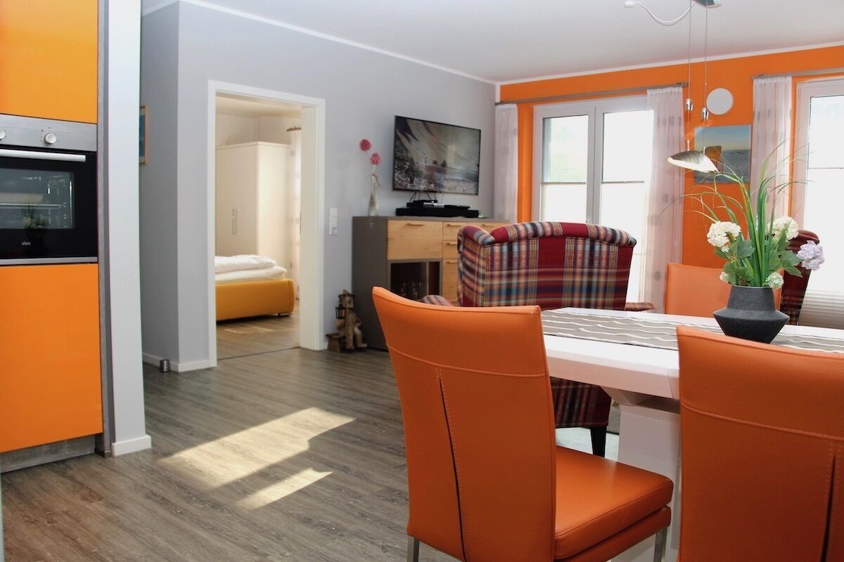 A modern kitchen with orange cabinetry, a dining area with orange chairs, and a television on the wall.