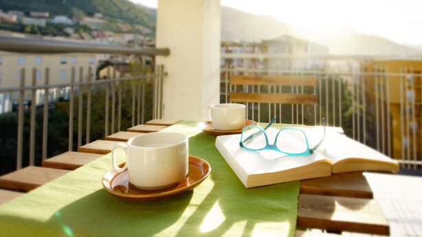 Ein Balkon mit einem Tisch, gedeckt für zwei Personen, mit Tasse, Untertasse und einem offenen Buch mit Gläsern darauf.