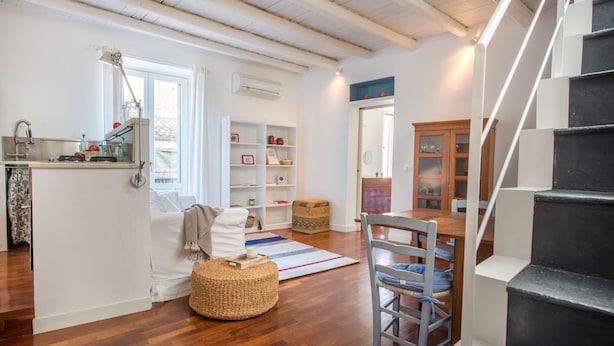 A spacious living area with a wooden floor, a white sofa, a wicker ottoman, and a dining table with chairs. A staircase with black steps leads to another level. - Terrazza al Capo by Wonderful Italy (Palermo)
