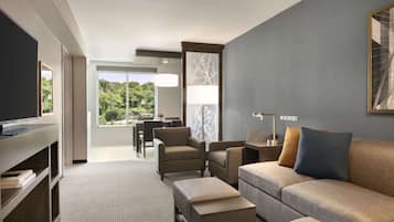 Una habitación de hotel moderna con sofá, sillones, televisor y zona de comedor con vistas a zonas verdes.