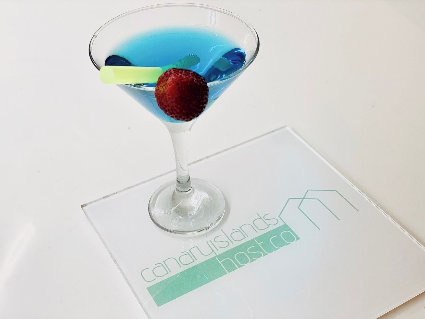 Een martini glas met een blauwe vloeistof en een aardbei op een wit oppervlak, met een logo met de tekst 
