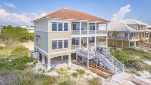 Une maison à deux étages située en bord de mer, dotée d’un balcon et d’un escalier menant à l’étage supérieur, avec une plage de sable en arrière-plan.