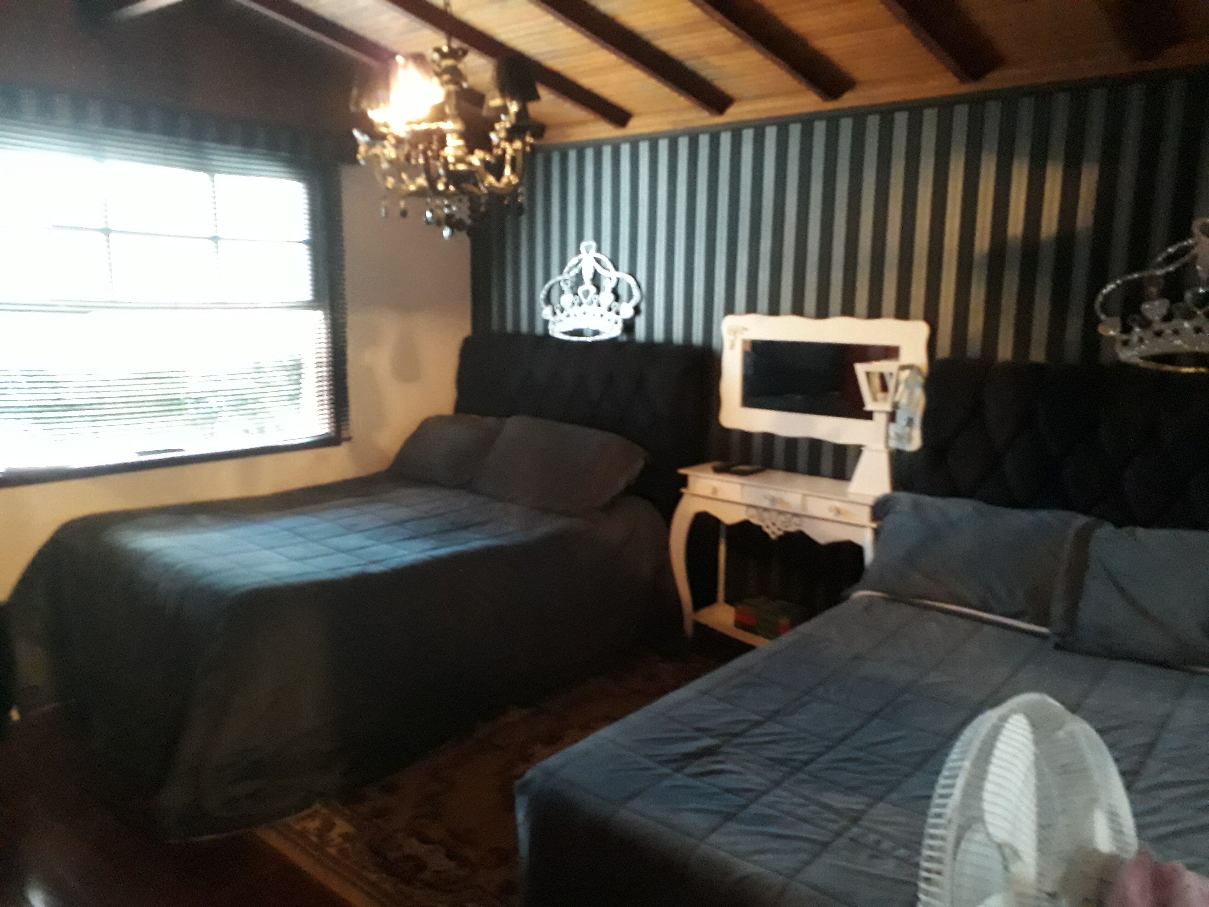 A room with two beds, a nightstand with a TV, a chandelier, and a window with blinds.