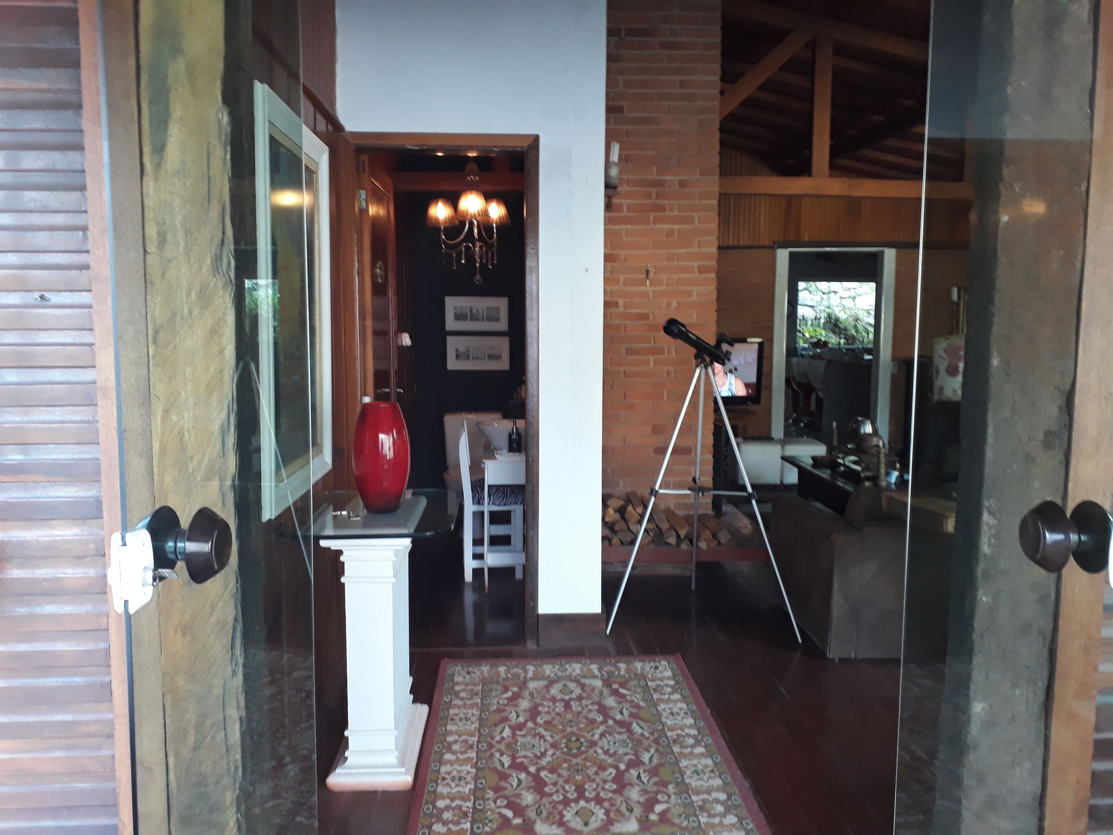 A modern interior with a glass door entrance, a red vase on a pedestal, a tripod with a telescope, and a patterned rug.