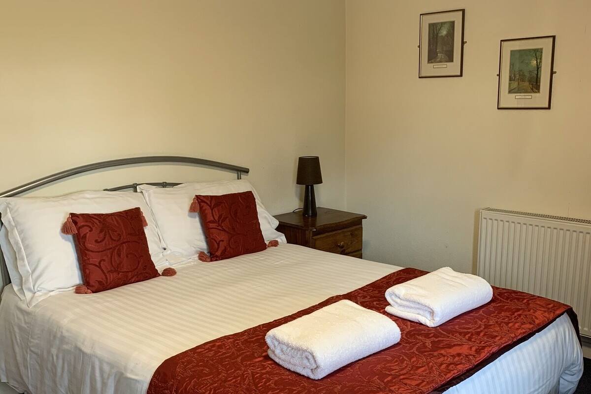 Un lit bien fait, avec du linge de lit blanc et rouge, une lampe de chevet et deux tableaux encadrés accrochés au mur.