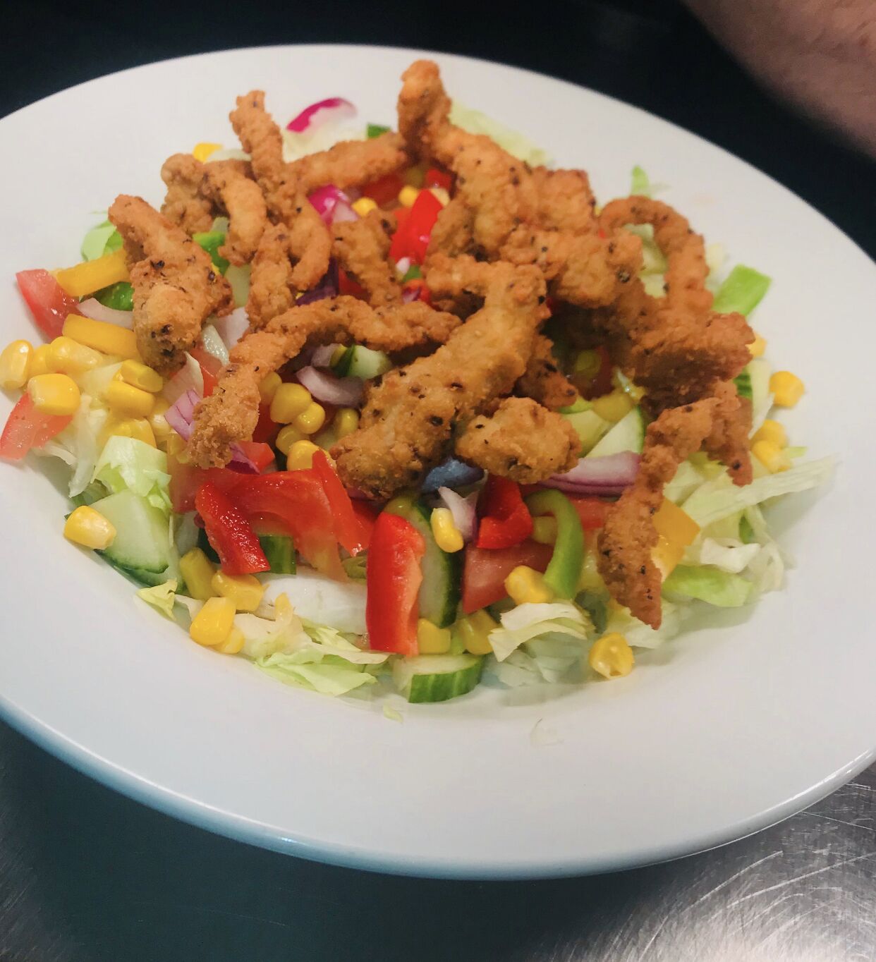 Ensalada con maíz, lechuga y pimientos rojos, coronada con trozos de pollo frito.