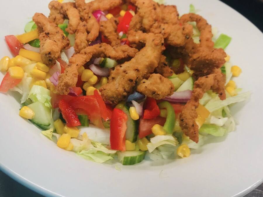 Ensalada con maíz, lechuga y pimientos rojos, coronada con trozos de pollo frito.