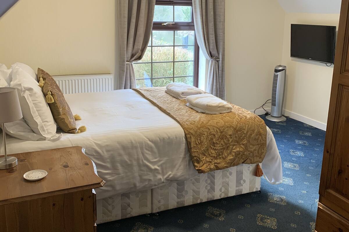 Une chambre à coucher avec un grand lit, une table de chevet, un téléviseur fixé au mur et une fenêtre avec des rideaux.