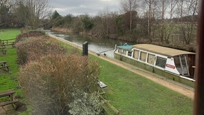 Ein Fluss mit einem vor Anker liegenden Hausboot namens 'Polly'. Es gibt Picknicktische und eine Rasenfläche mit Büschen.