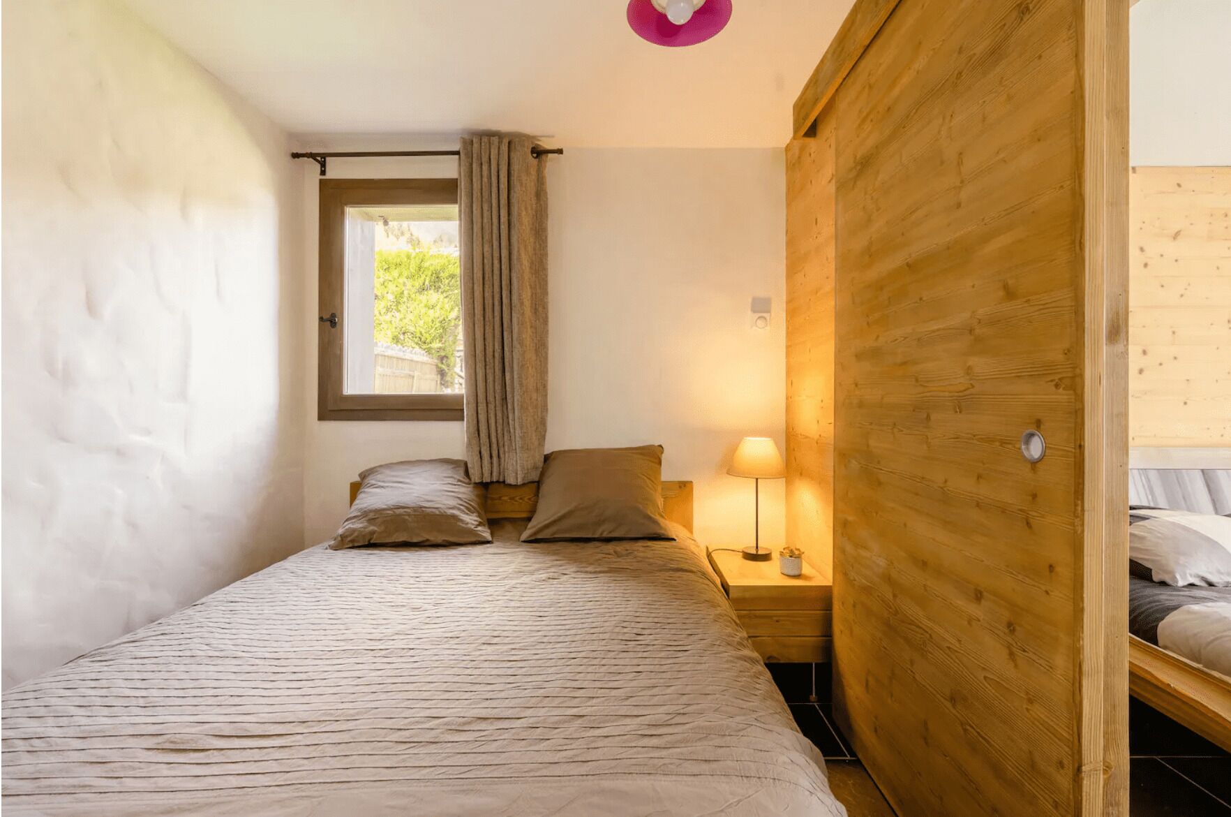 Un dormitorio pequeño y acogedor con una cama individual, una mesita de noche con lámpara, un armario de madera y una ventana con cortinas.