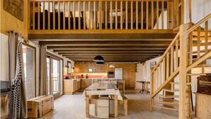 Een ruime loftkamer met een hoge plafond, zichtbare houten balken, een eethoek met een lange tafel, en een keuken met houten kastjes en een groot raam.