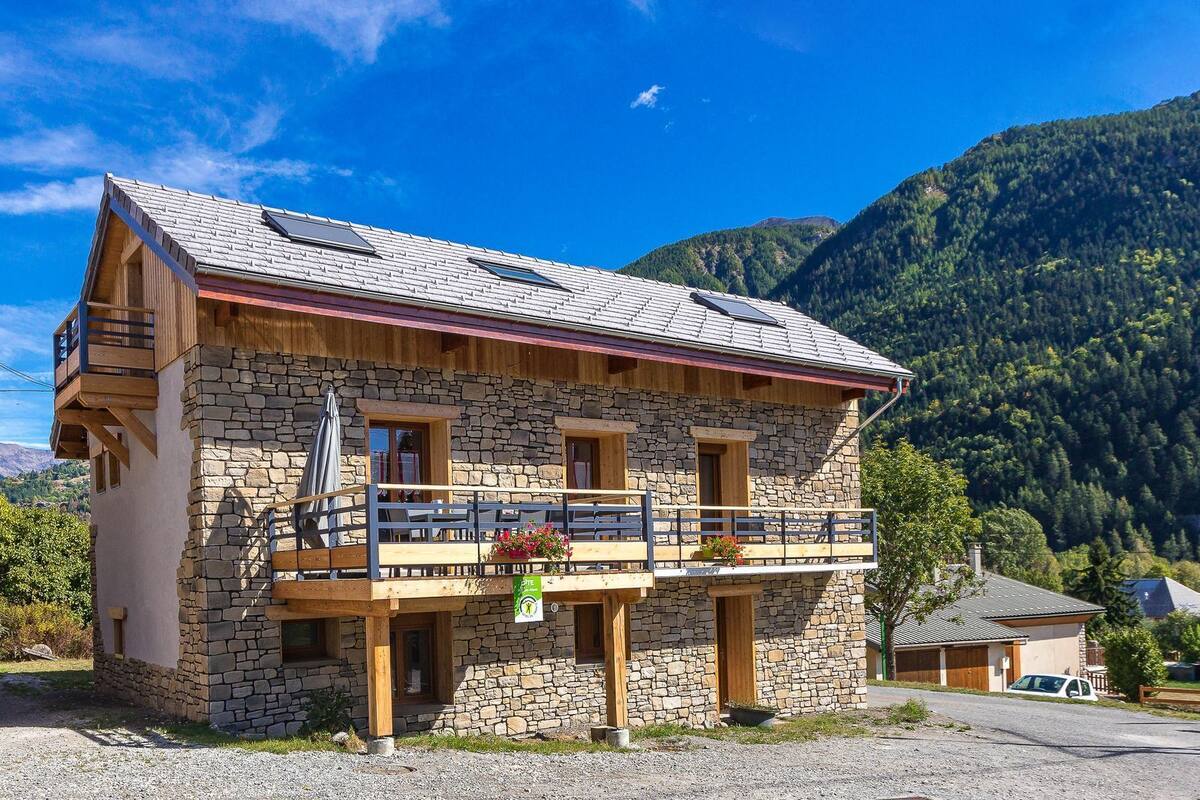 Un edificio de piedra con balcón, terraza de madera y montañas al fondo.