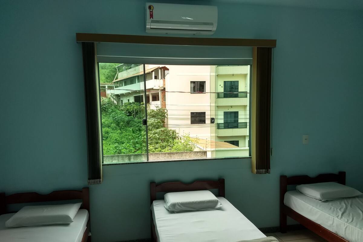 A room with two single beds, a window with a view of buildings and greenery, and an air conditioning unit above the window.
