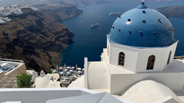 Un bâtiment blanc surmonté d’un dôme bleu, qui surplombe une zone côtière avec des bateaux et des falaises.