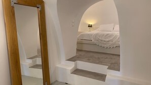 Une chambre avec un lit blanc, une table de chevet et une lampe. On y trouve un cadre de porte en bois et un escalier blanc menant au lit.