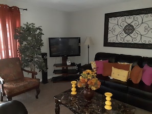 Uma sala de estar com televisão, sofá, poltrona, planta e um vaso com flores.