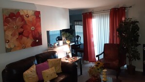 A living room with a dark leather sofa, a coffee table, and a large floral painting on the wall. There is a dining area in the background with a table and chairs. - Master's apartment rental (Augusta)