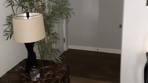 A hallway with a dark brown floor, a white door, a framed artwork, a potted plant, and a lamp on a wooden stool. - Master's apartment rental (Augusta)