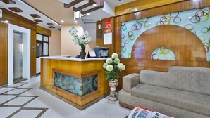 A reception area with a wooden reception desk, a sofa, a vase with flowers, and a wall with circular patterns. - Hotel Merit (Surat)