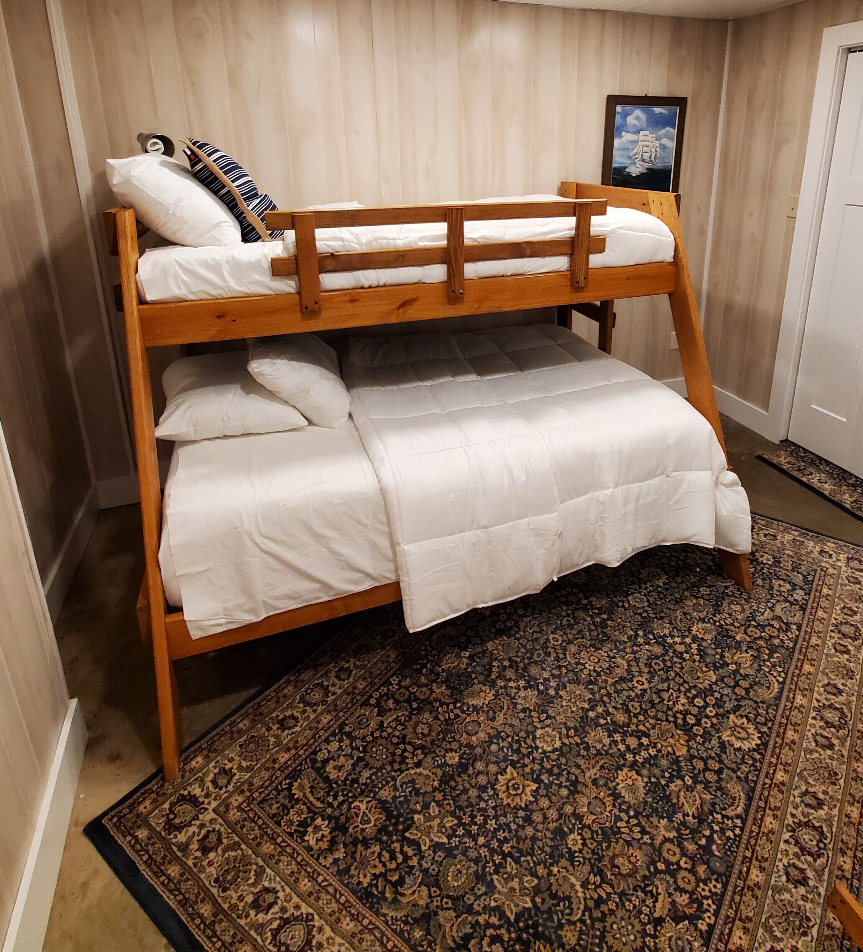 Un lit superposé avec une literie blanche, un tapis à motifs et un tableau encadré accroché au mur.