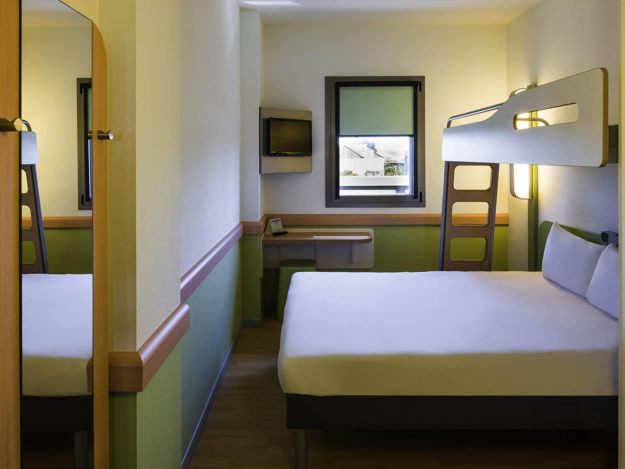 A compact hotel room with a bed, a desk, a mirror, a television, and a window with a view of buildings.