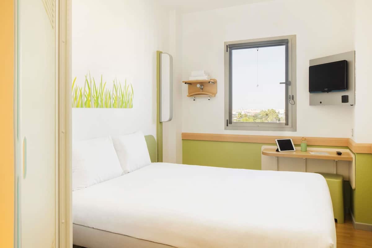 A hotel room with a bed, a desk with a computer, a TV, a window, and a wall-mounted shelf.