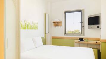Habitación de hotel con cama, escritorio con computadora, televisión, ventana y estante fijado a la pared.